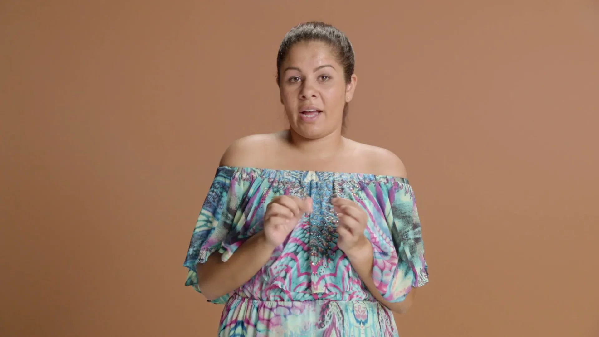 A woman in her 30s, wearing a colorful off-the-shoulder dress, stands against a brown background, speaking directly to the camera.