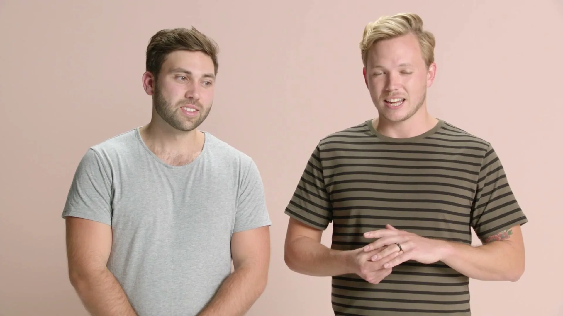 Two men, one in a grey t-shirt and one in a striped t-shirt, stand side-by-side, talking and gesturing with their hands in a studio.