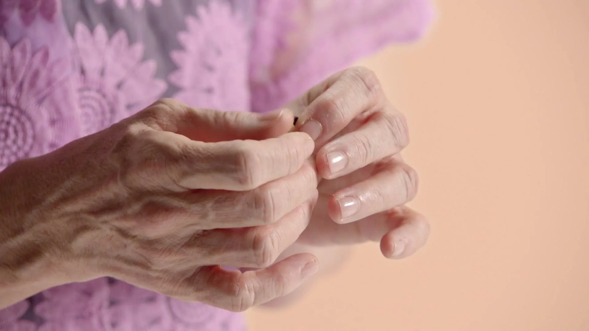 An older woman's hands, with painted nails, delicately hold and manipulate a small, dark object against a soft pink background.