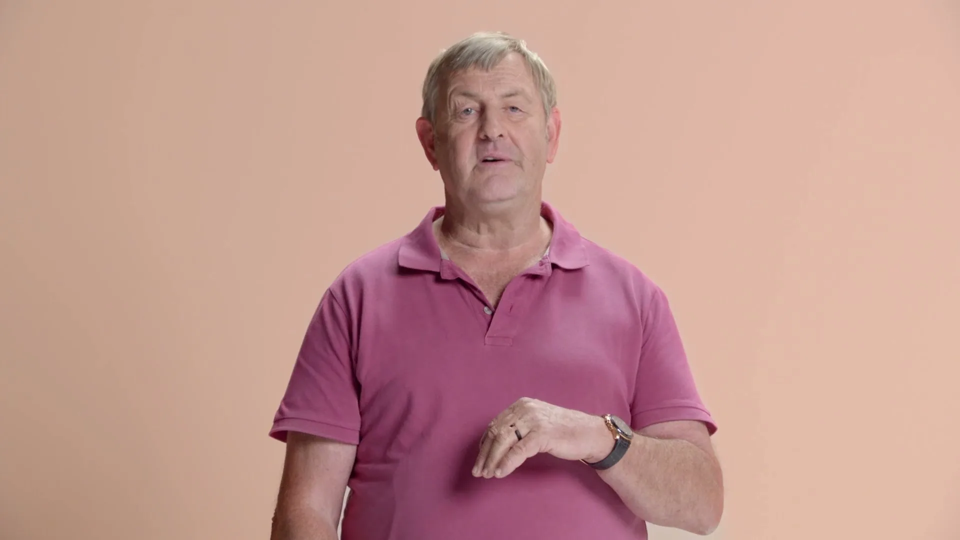 A man in his 60s, wearing a pink polo shirt, stands against a peach background, making a hand gesture.
