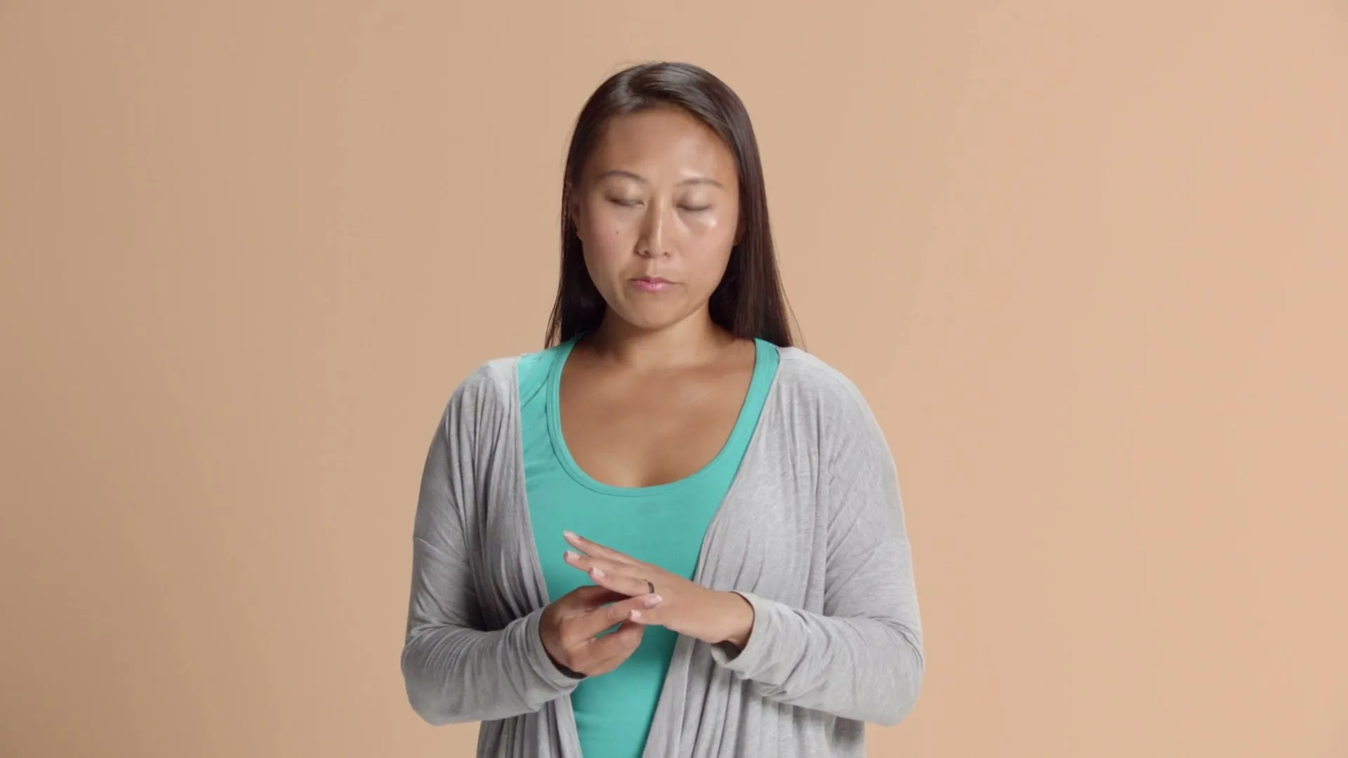 A young Asian woman in a teal top and grey cardigan looks down at her hands, then up at the camera, against a peach background.