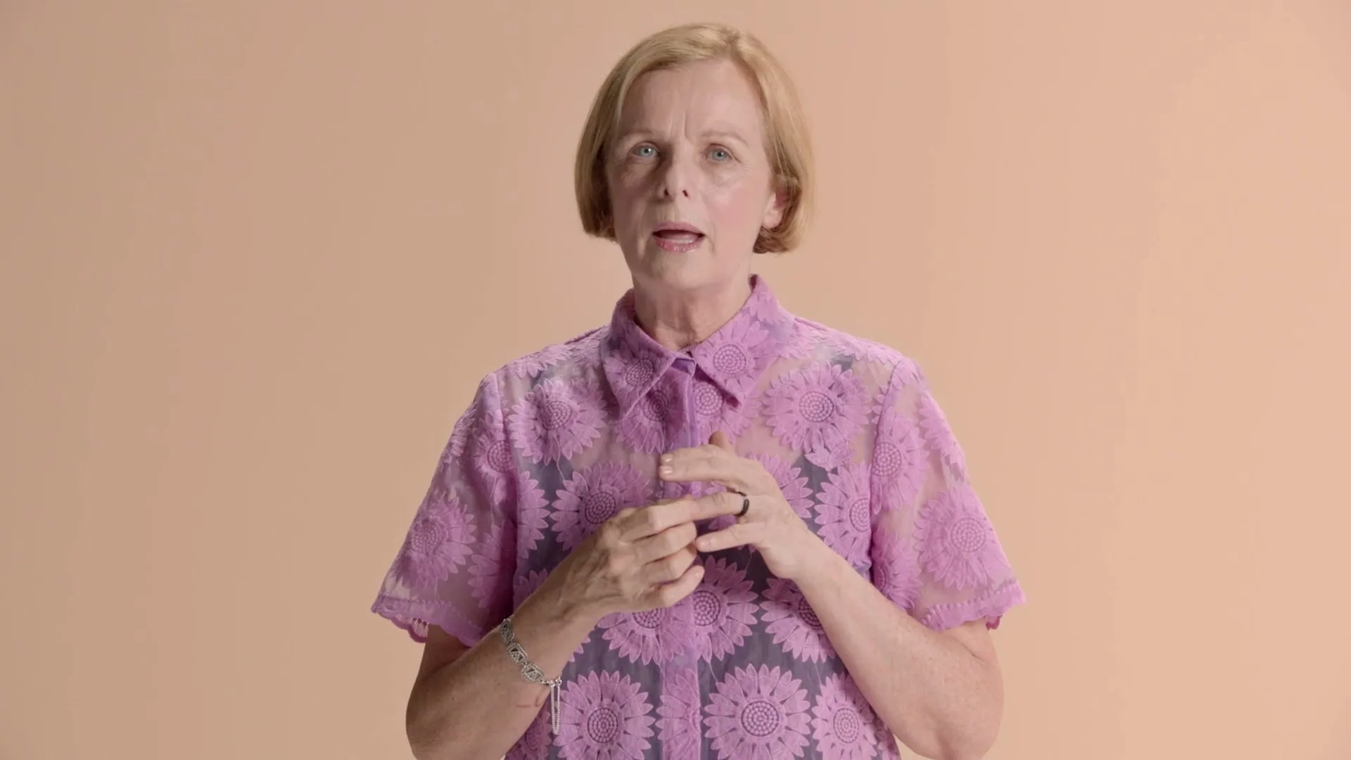 A blonde woman in her 60s, wearing a purple floral shirt, stands against a peach background, looking directly at the camera.