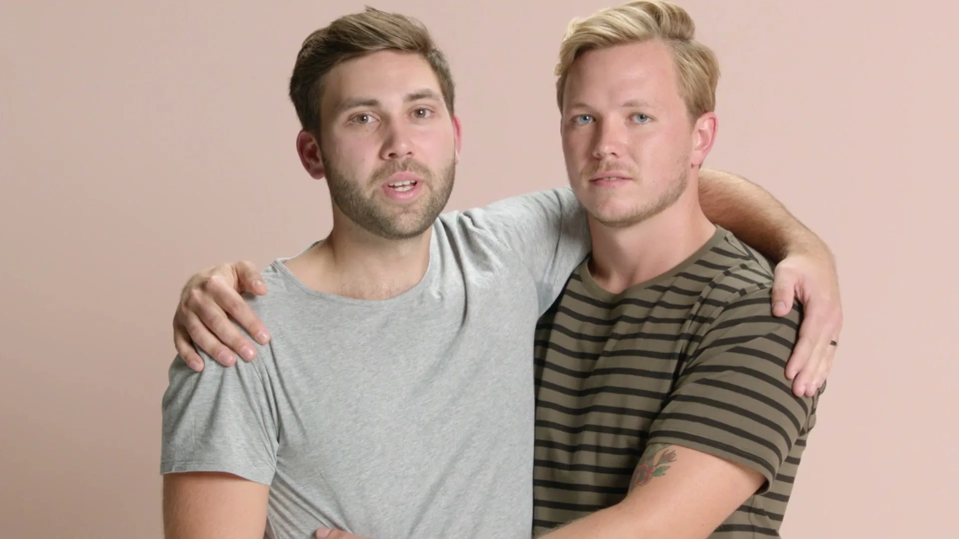 Two men, one with a beard and one with blonde hair, stand embraced against a plain pink background, looking directly at the camera.