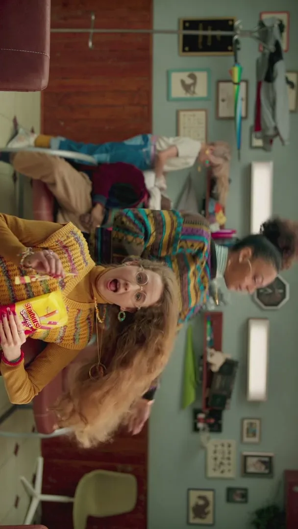 A young woman with long, curly hair holds a bag of Starburst Swirlers and eats one, while her hair is being styled by a hairdresser in a salon.
