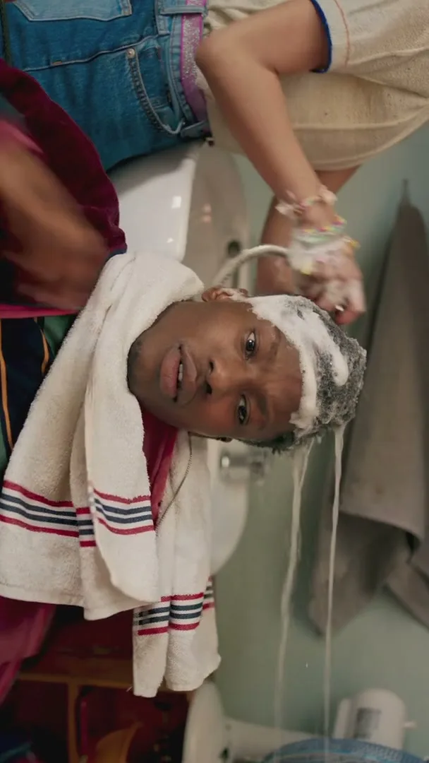 A young man with a towel around his neck is getting his hair washed in a sink by another person, in a bathroom.