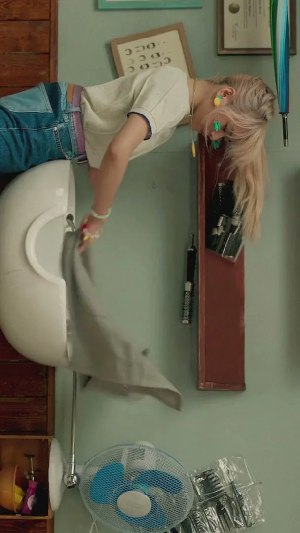 A young woman with blonde hair and colorful clothes is upside down, washing her hair in a salon sink, with a fan and hair products visible.