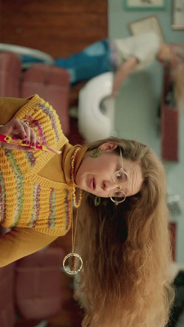 A woman with long, curly hair and glasses, wearing a yellow knitted vest, holds a Starburst Swirler and looks surprised in a living room.