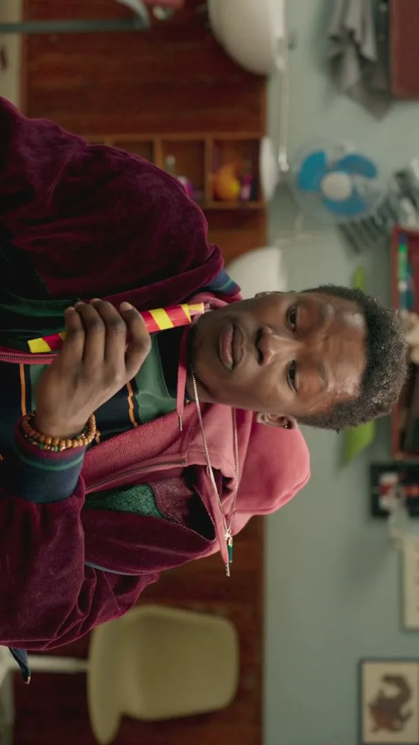 A young Black man in a maroon robe and pink hoodie holds a Starburst Swirler candy to his mouth, looking surprised in a messy room.
