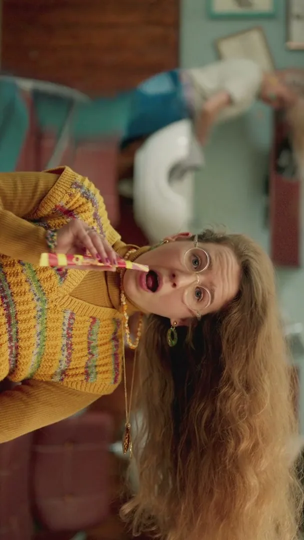 A woman with long, curly hair and glasses, wearing a yellow knitted vest, holds a Starburst Swirler candy in her mouth, looking surprised in a living room.