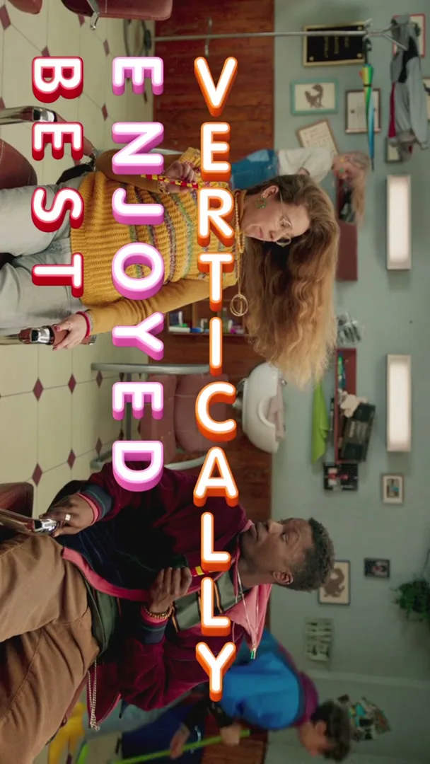A young man and woman are sitting in a barbershop, looking at the camera, with the text "BEST ENJOYED VERTICALLY" superimposed over them.