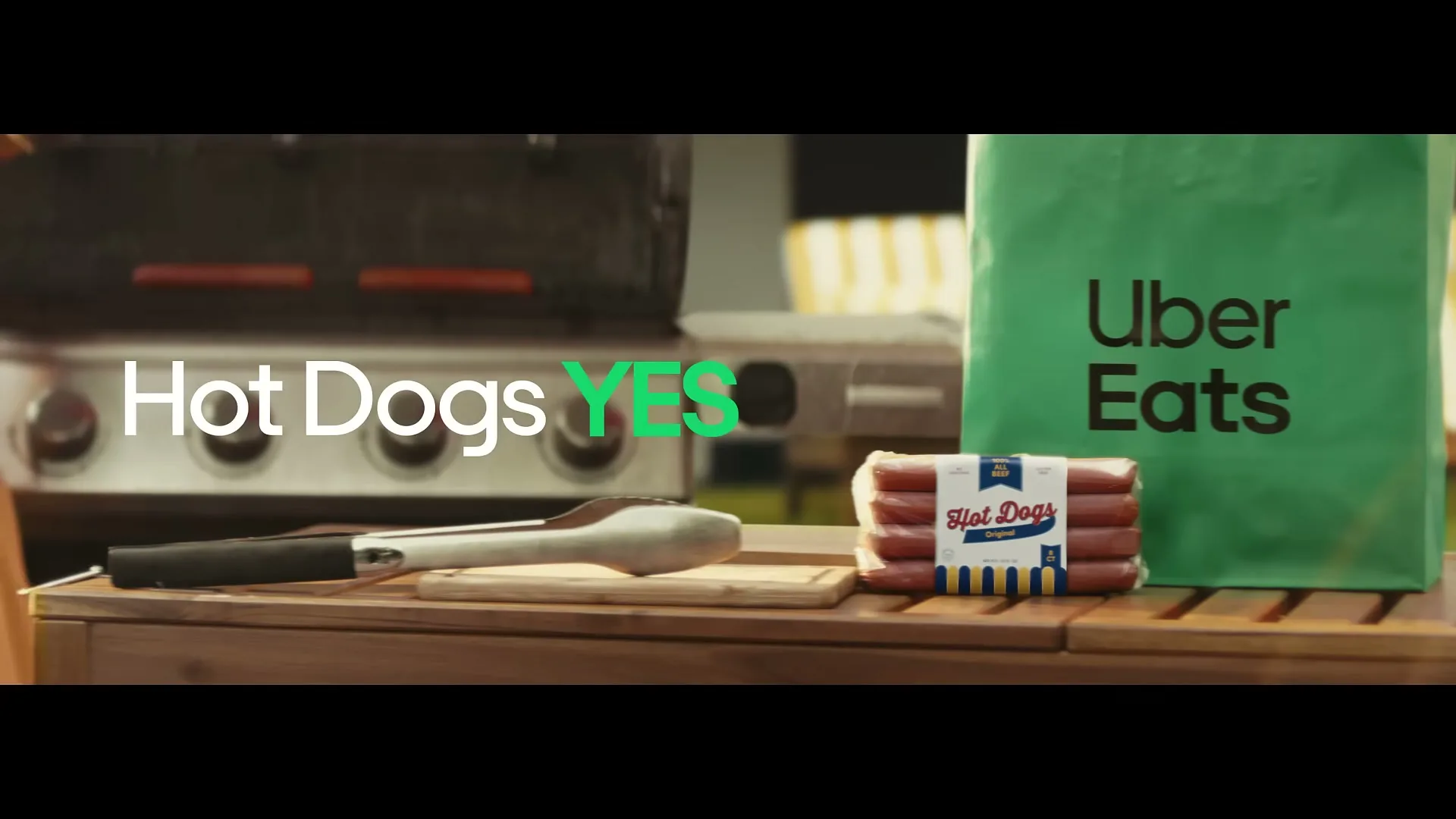 A package of hot dogs and an Uber Eats bag sit on a wooden table next to a barbecue grill, with "Hot Dogs YES" text on screen.