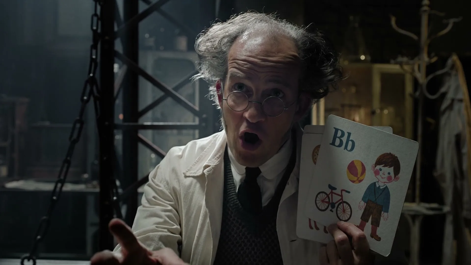 A male scientist in a lab coat and glasses holds up a flashcard with the letter 'B' and a boy on it, speaking in a dimly lit laboratory.