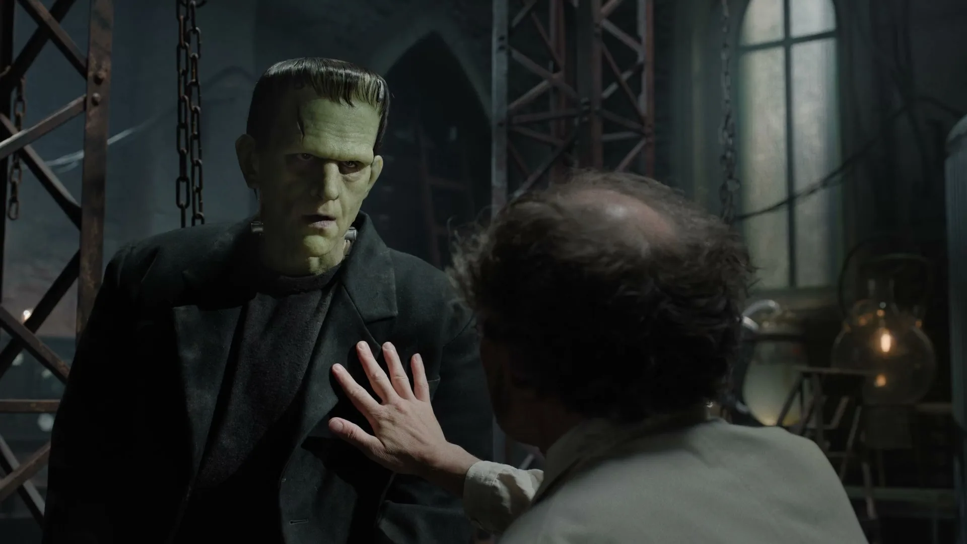 Frankenstein's monster, green-skinned and in a dark suit, stands as a man in a lab coat reaches out to touch his chest in a dimly lit laboratory.