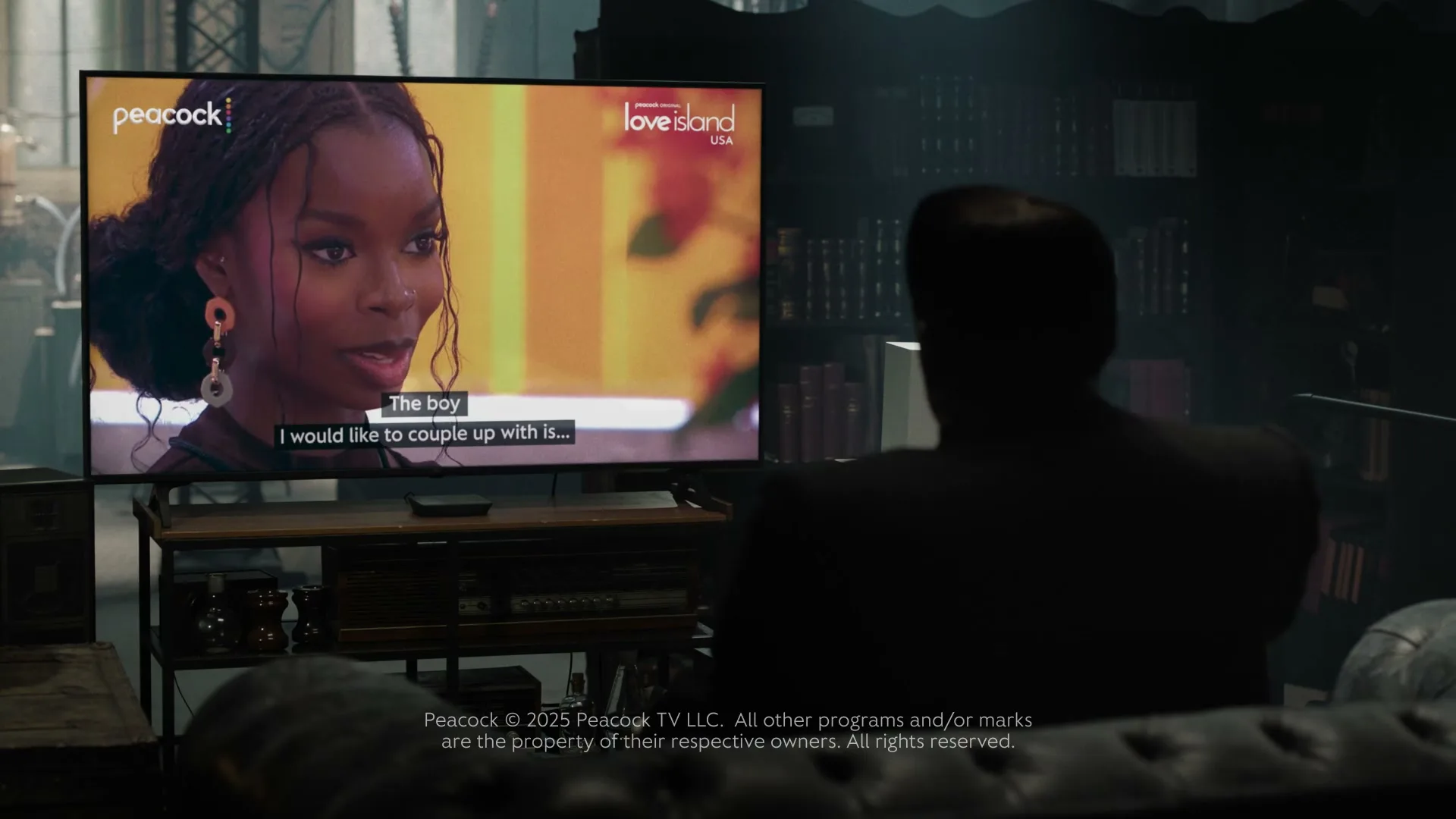 A man in silhouette watches a woman on a TV screen, who is speaking with subtitles, in a dimly lit room.