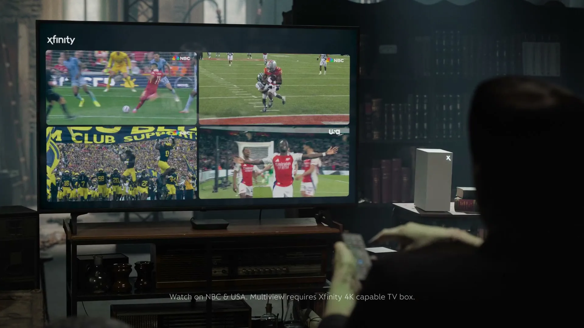 A person with green skin and dark hair watches four sports games simultaneously on a large TV in a dimly lit, cluttered room.