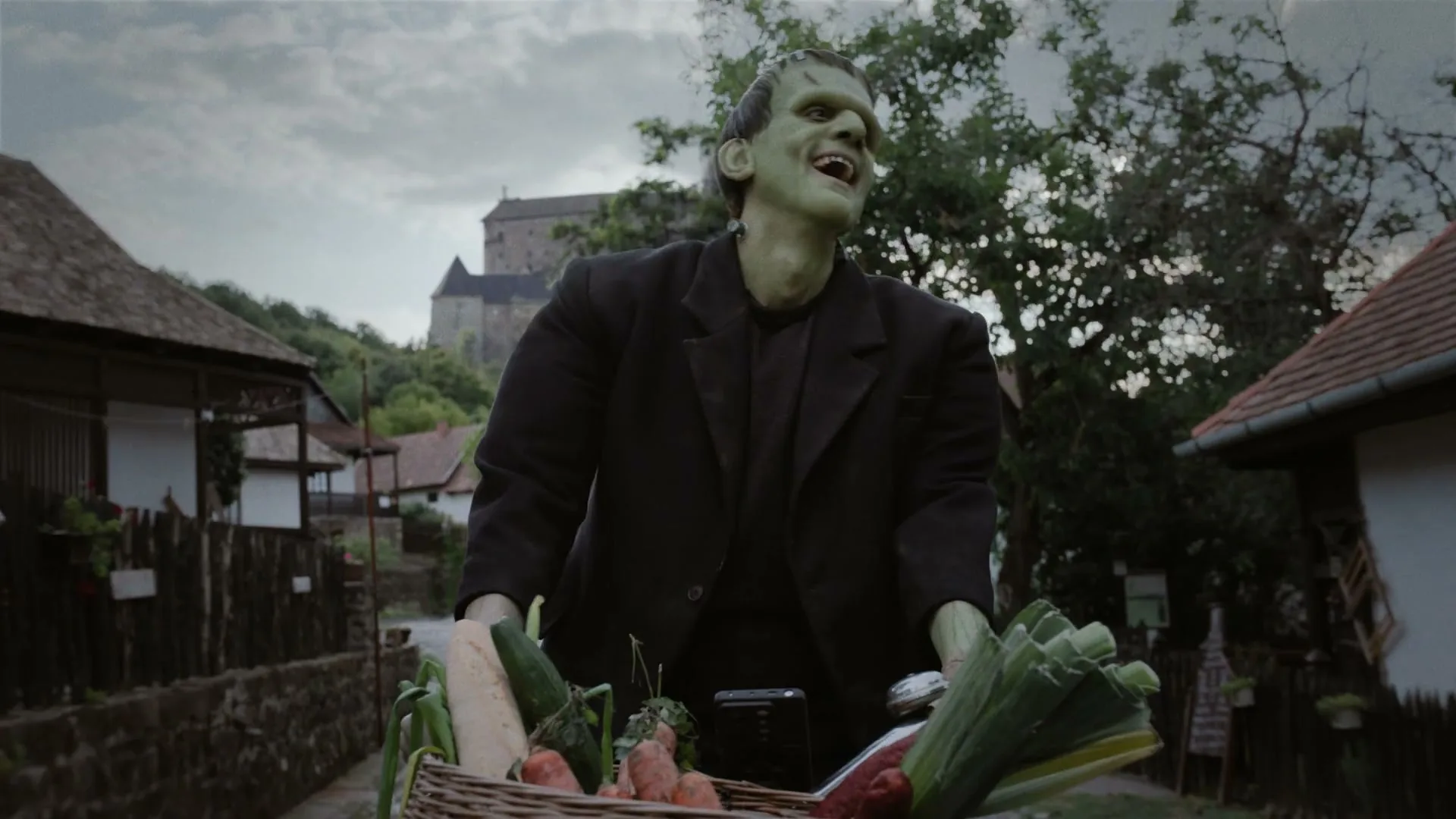 Frankenstein's monster, with green skin and a dark suit, rides a bicycle with a basket of groceries through a village street.