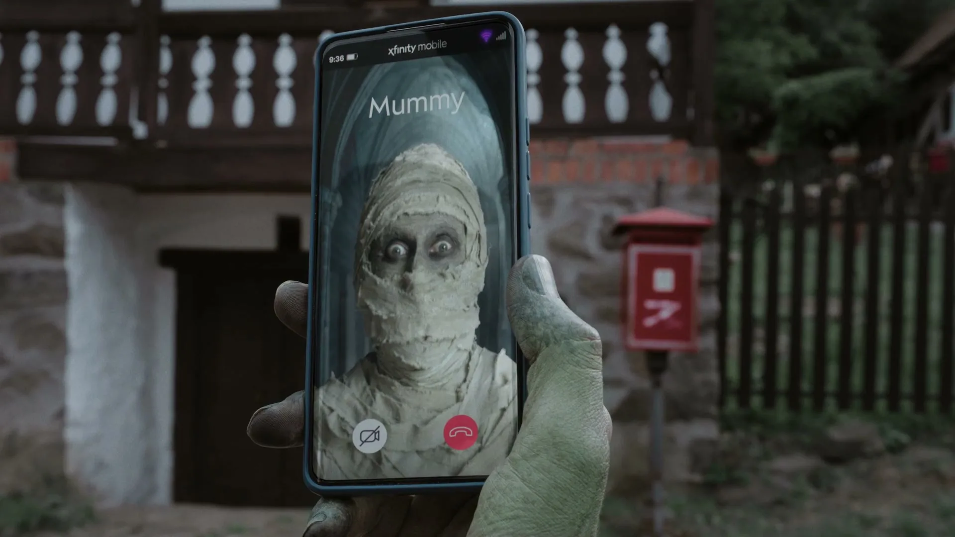A green, monstrous hand holds a smartphone displaying a video call from a mummy, with a rustic house and red mailbox in the background.