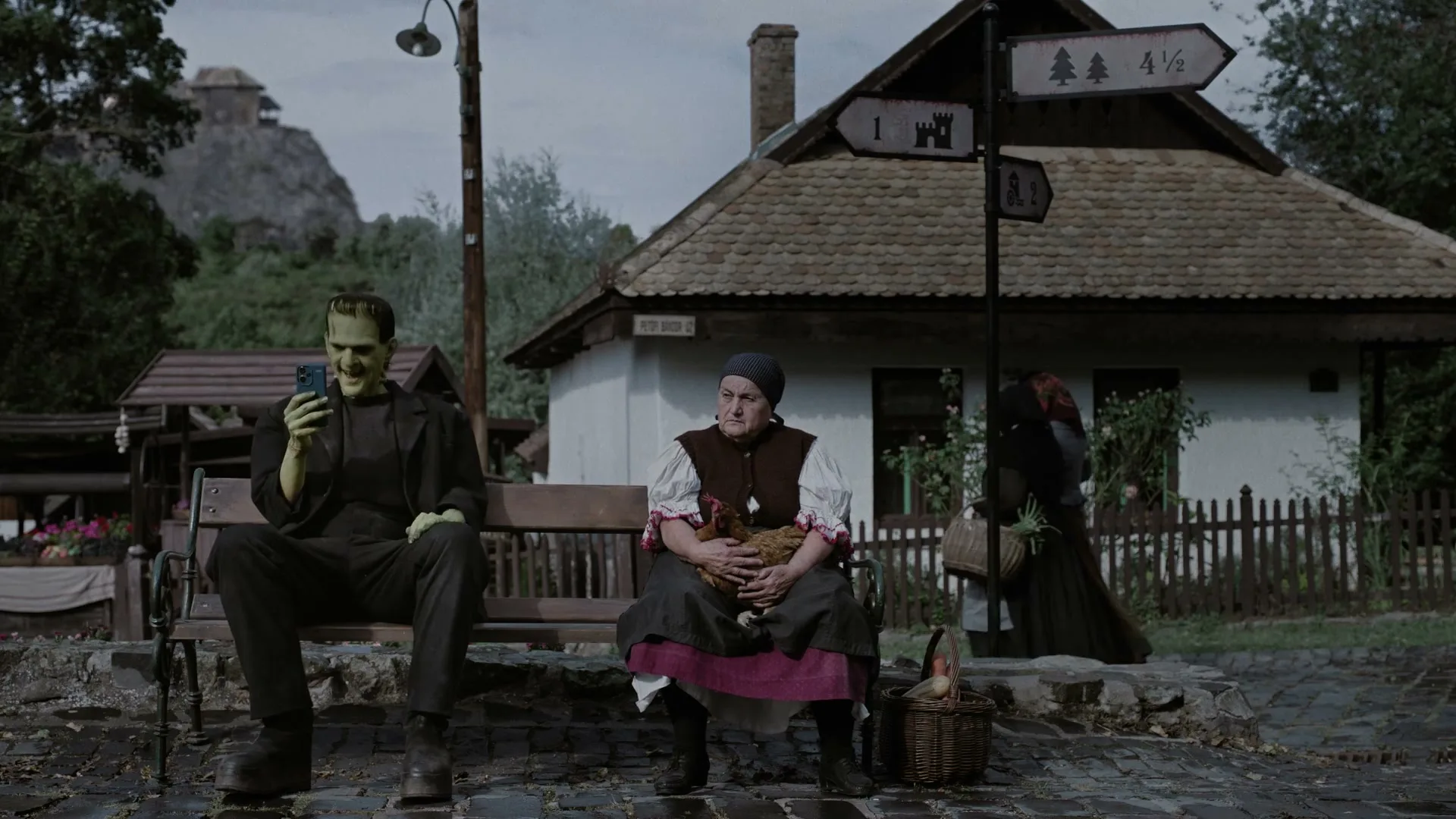 Frankenstein, a green-skinned man in a dark suit, sits on a bench looking at his phone next to a woman holding a chicken in a rural village.