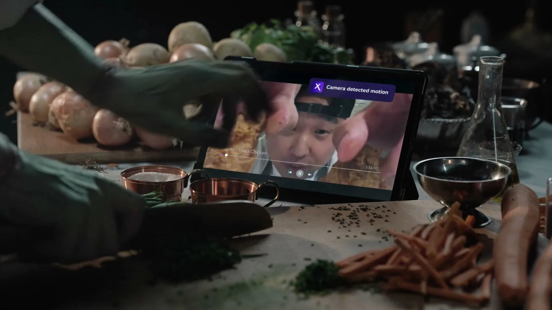 A person with green skin chops herbs on a cutting board while watching a cooking video on a tablet, which then switches to a security camera feed.