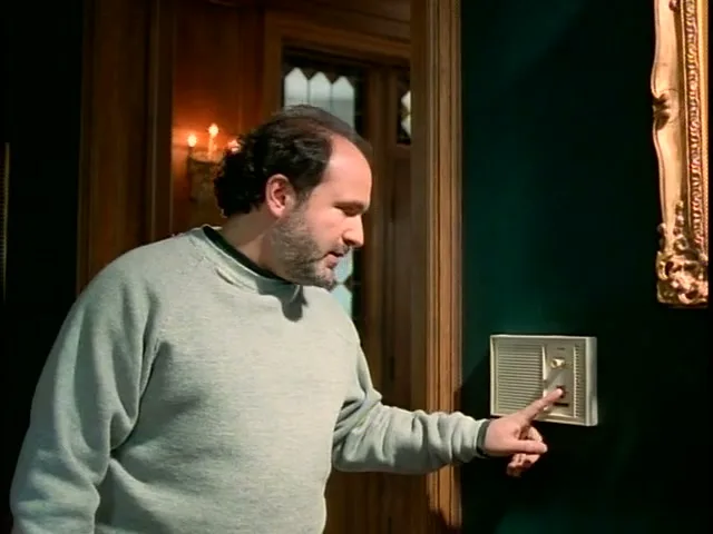 A man in a grey sweatshirt adjusts a thermostat on a dark green wall in a large, ornate house, looking confused.