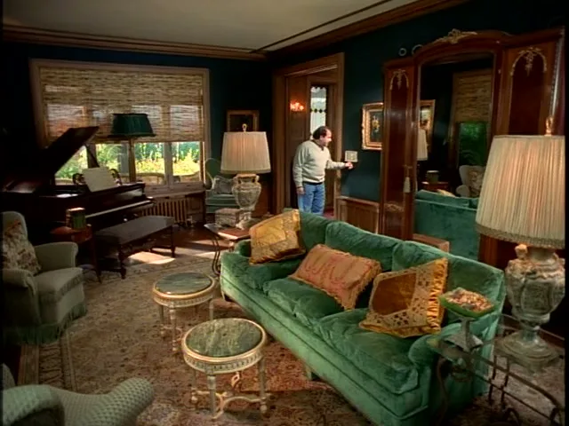 A man in a white sweater and blue jeans stands in a doorway of a luxurious living room with a green couch and a grand piano.