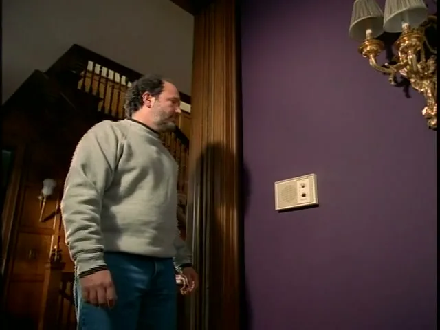 A man in a grey sweatshirt and jeans walks into a large house and presses a button on an intercom on a purple wall.