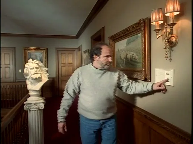 A man in a grey sweatshirt and jeans walks down a hallway in a large house, then stops to press a button on an intercom.