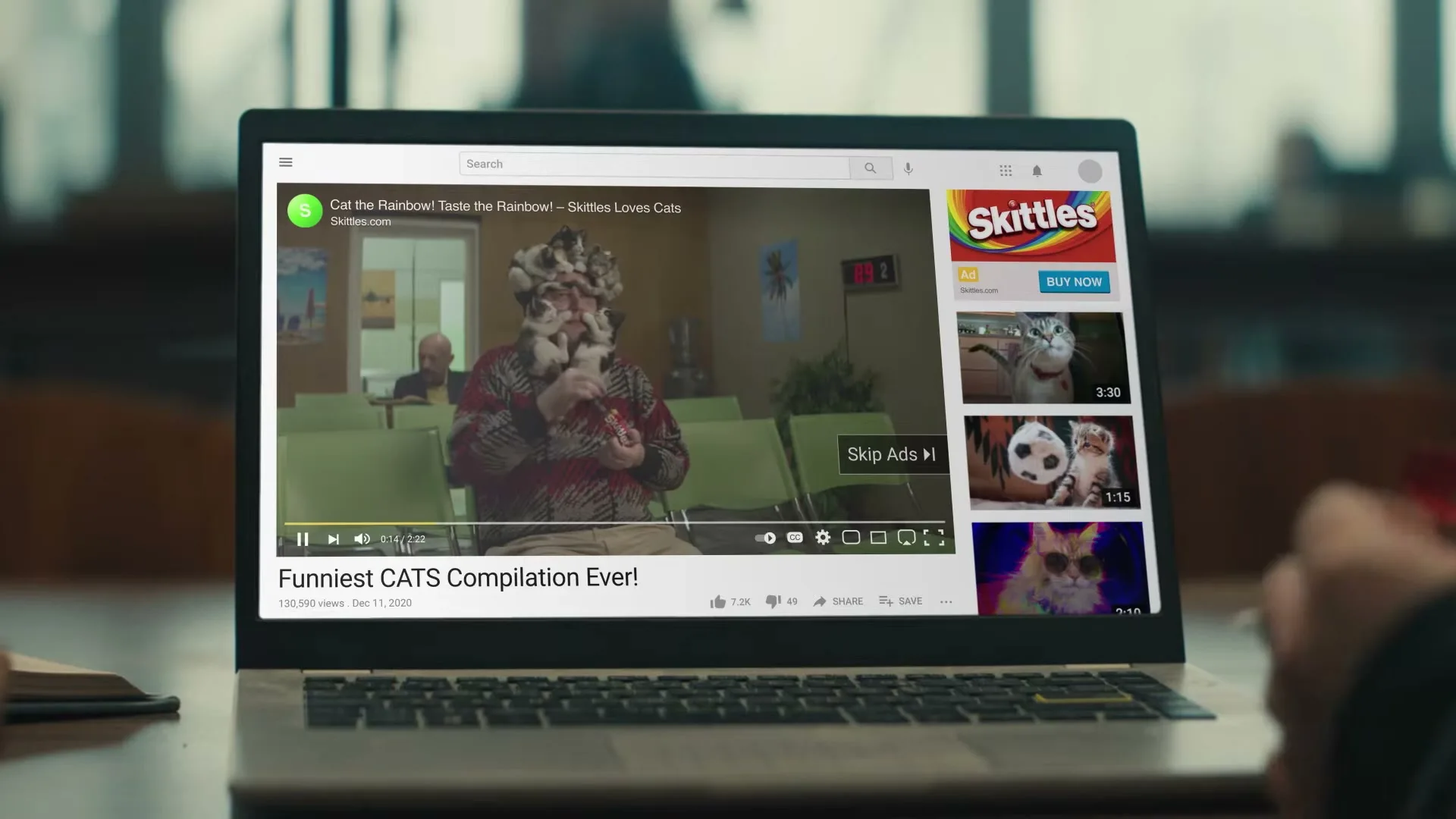 A laptop screen displays a YouTube video of a man covered in cats, with a Skittles ad playing on the side.