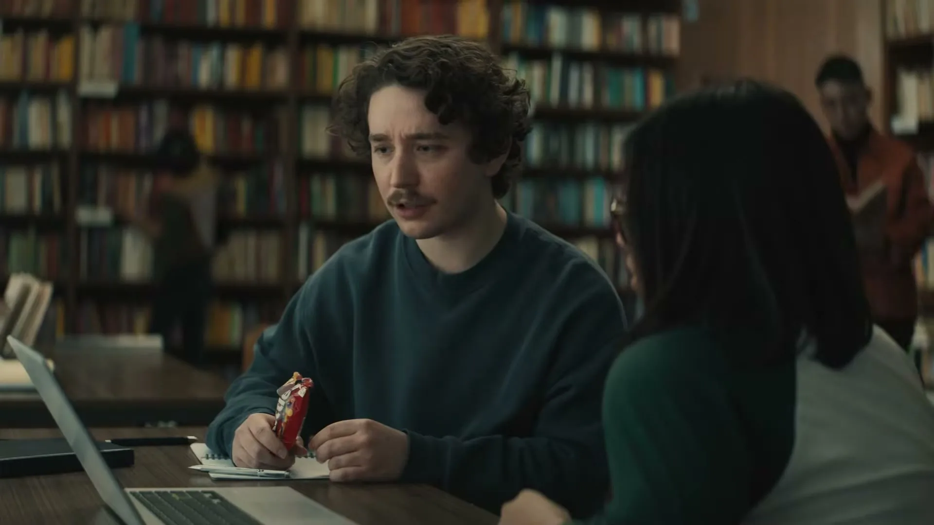 A young man with curly hair and a mustache sits at a library table, holding a Skittles package and talking to a woman.