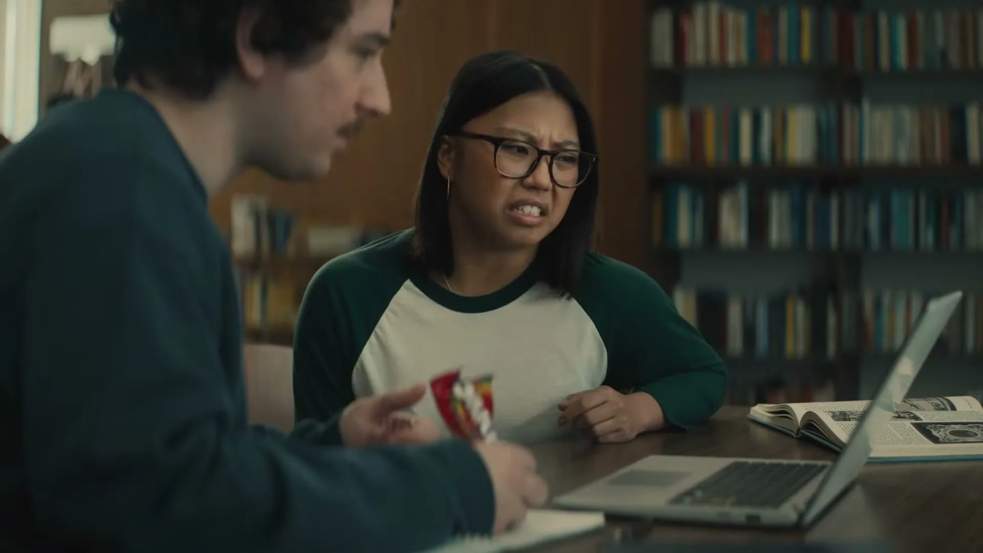 A young woman with glasses and a young man are sitting at a table in a library, looking at a laptop and a bag of Skittles.