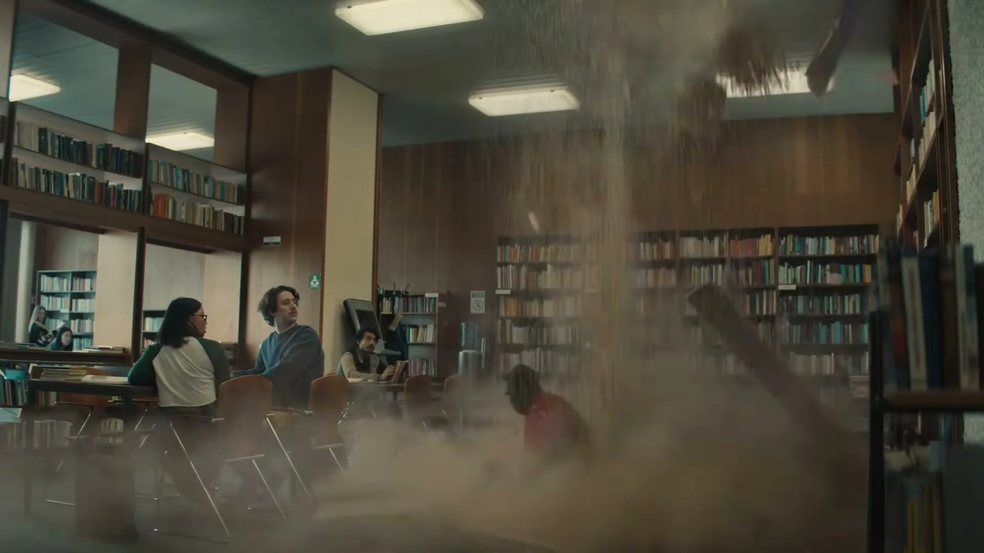 A man in a red shirt explodes into a cloud of dust and debris in a library, startling students.