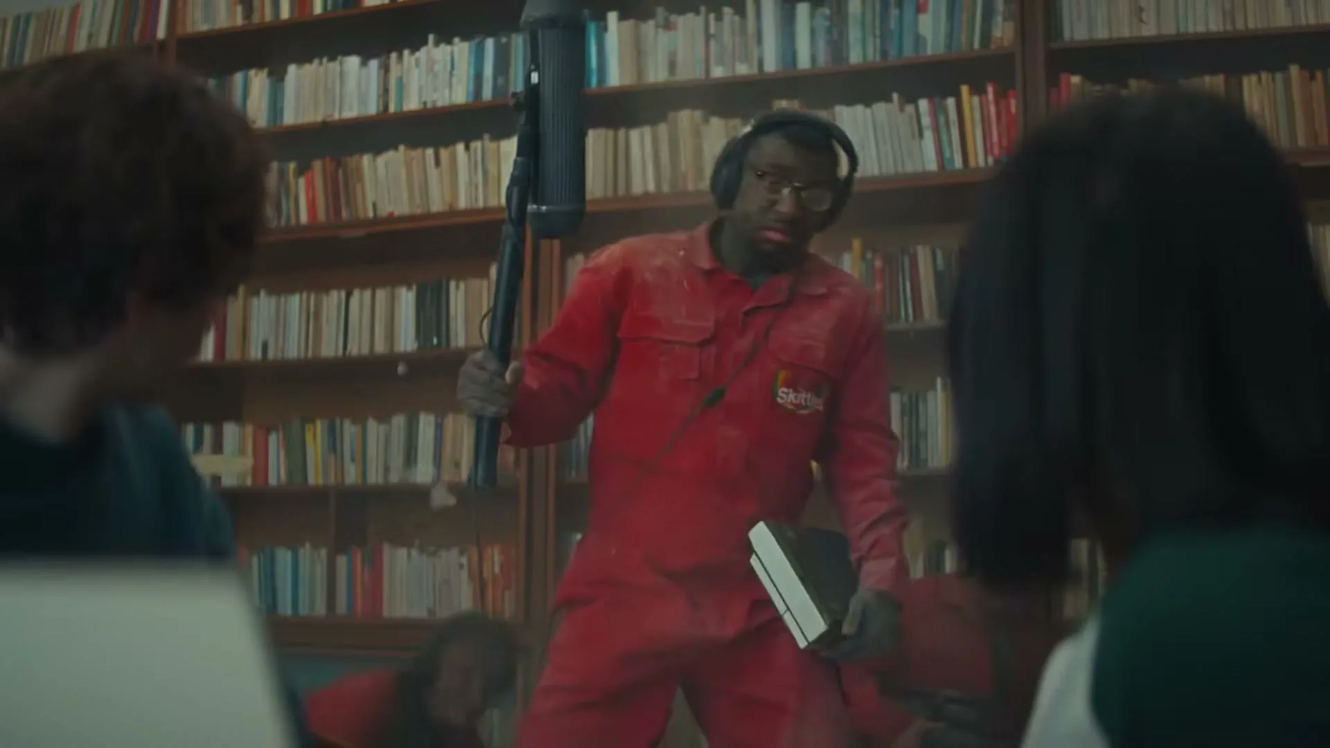 A man in a red jumpsuit and headphones, covered in dust, holds a boom mic and a book, looking surprised in a library.