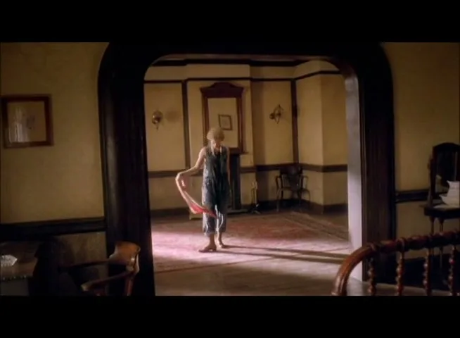 A young woman in denim overalls stands barefoot in a sunlit, empty room, twirling a pink scarf playfully.