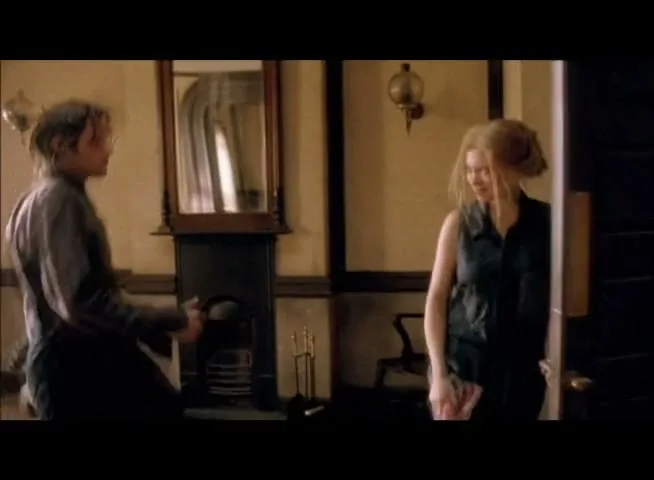A young man in dark clothes and a young woman in a denim dress are in a room, the man turns to the woman, who smiles.
