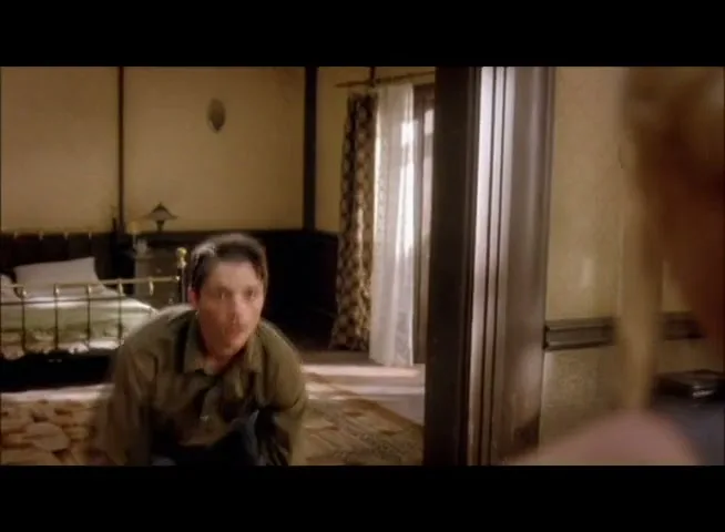 A man in a green shirt and dark pants looks up at a woman, who is out of frame, in a bedroom.