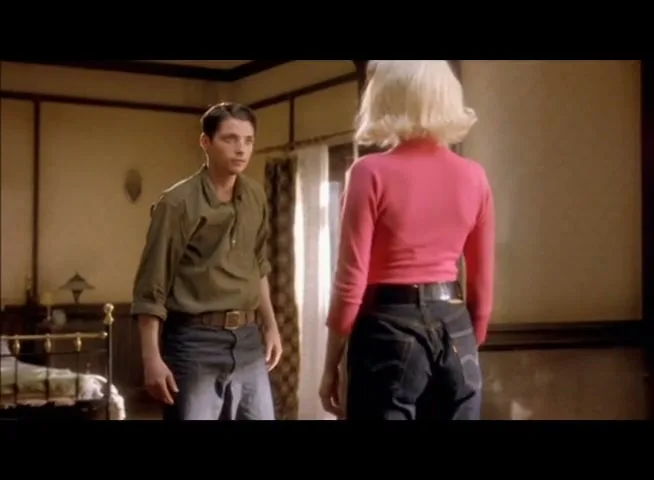 A young man in a green shirt and jeans looks at a blonde woman in a pink top and jeans in a dimly lit room.