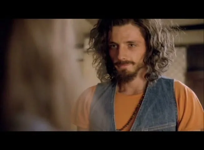 A young man with long, curly hair and a beard, wearing a denim vest and orange shirt, looks intently at someone off-screen, then leans in for a kiss.