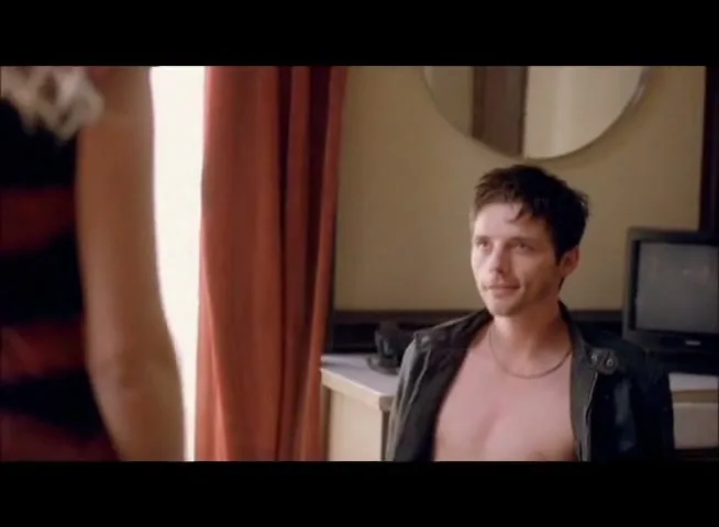 A young man with dark hair and an open leather jacket sits on a bed, looking up at a blonde woman whose back is to the camera in a hotel room.
