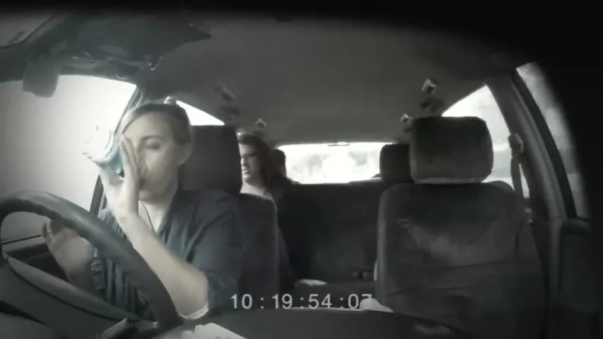A young woman in a car inhales from a plastic bag, while another person is visible in the back seat, as the car interior darkens.