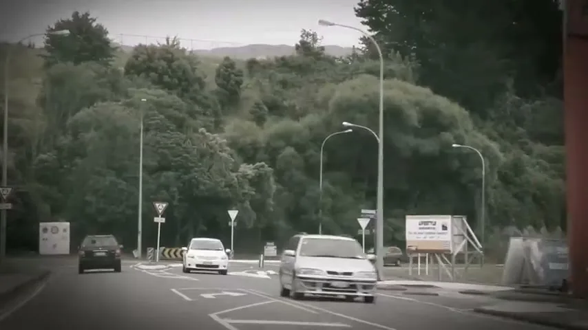 Cars drive on a road with a roundabout, surrounded by green hills and commercial buildings under an overcast sky.