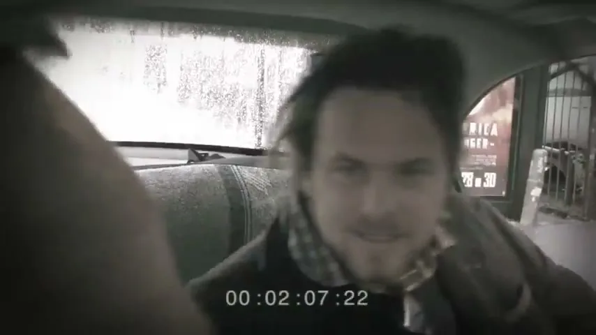A disheveled man with messy hair and a scarf looks back from the passenger seat of a car, with rain on the back window.