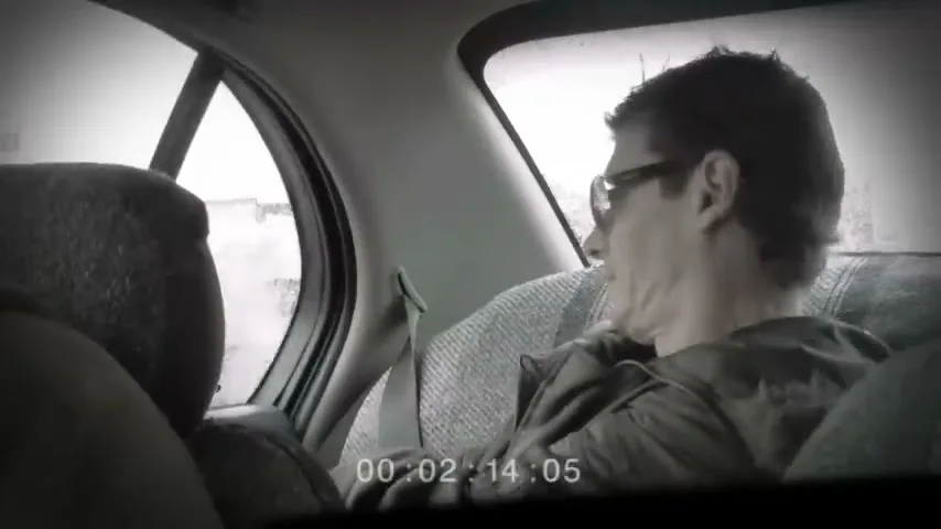 A man with glasses in the back seat of a car turns to look out the window, then turns to face forward, looking confused.