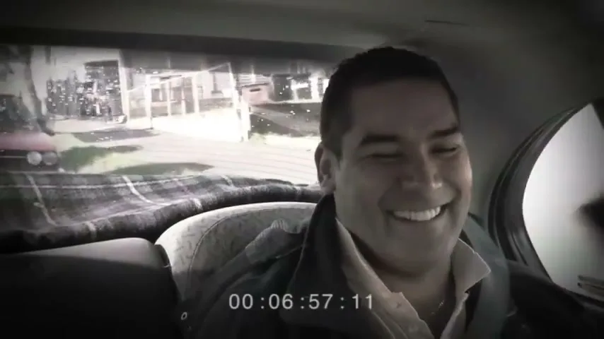 A man with dark hair and a light shirt laughs in the back seat of a car, with a blurry view of the outside world through the windows.