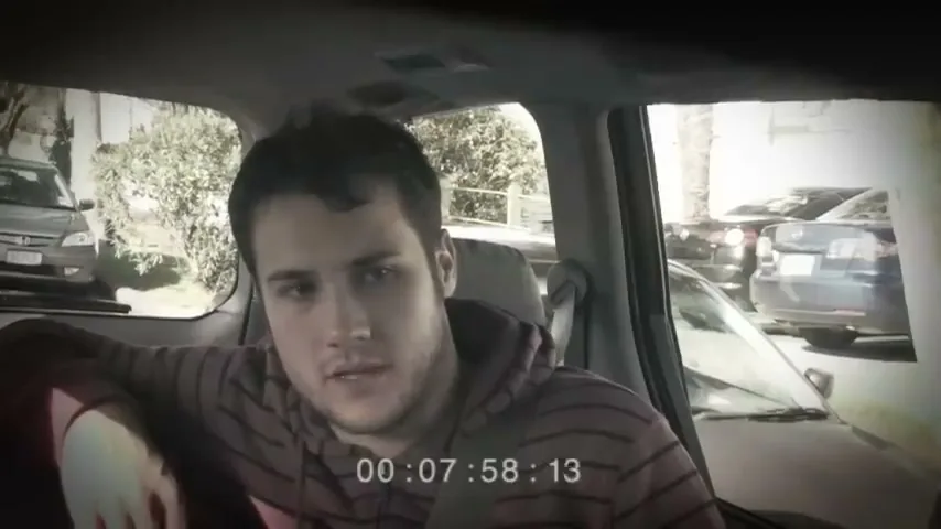A young man with dark hair and a striped hoodie sits in the passenger seat of a car, looking tired and disoriented, as cars are visible outside the windows.