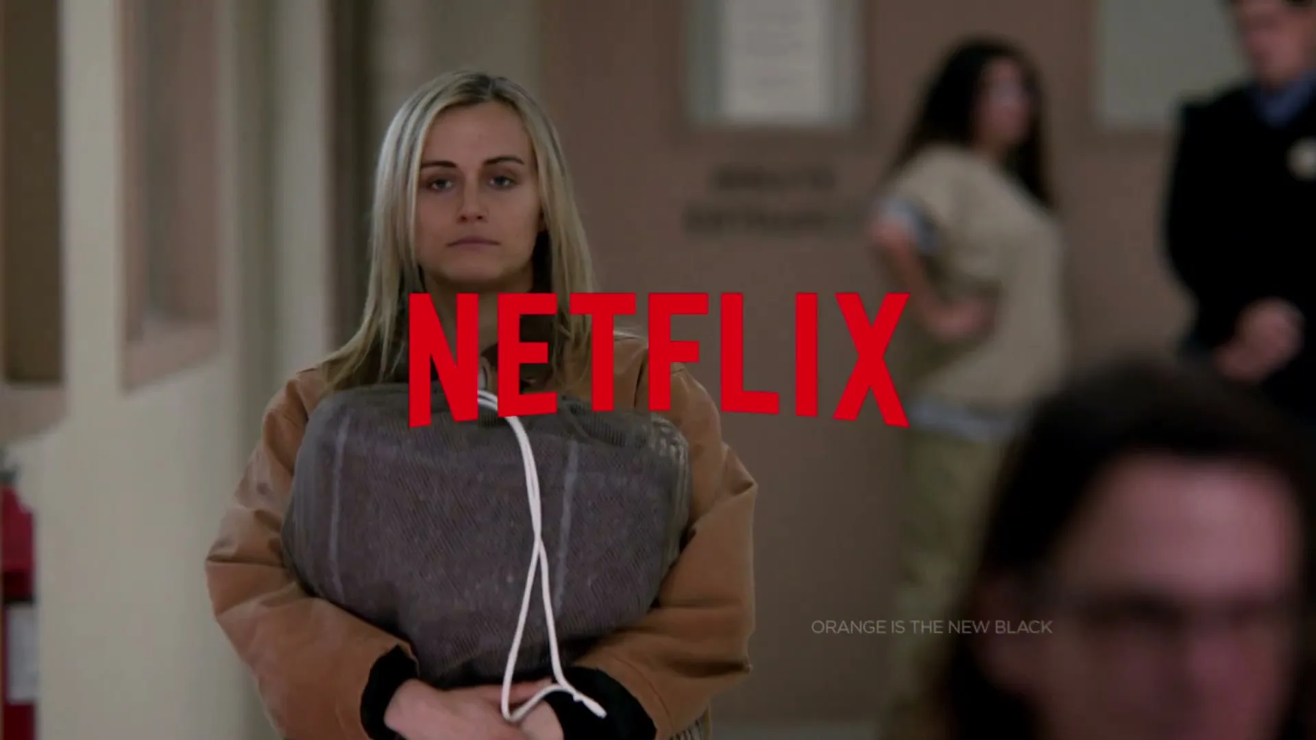 A blonde woman in an orange jumpsuit holds a bundled blanket, looking forward in a prison hallway.