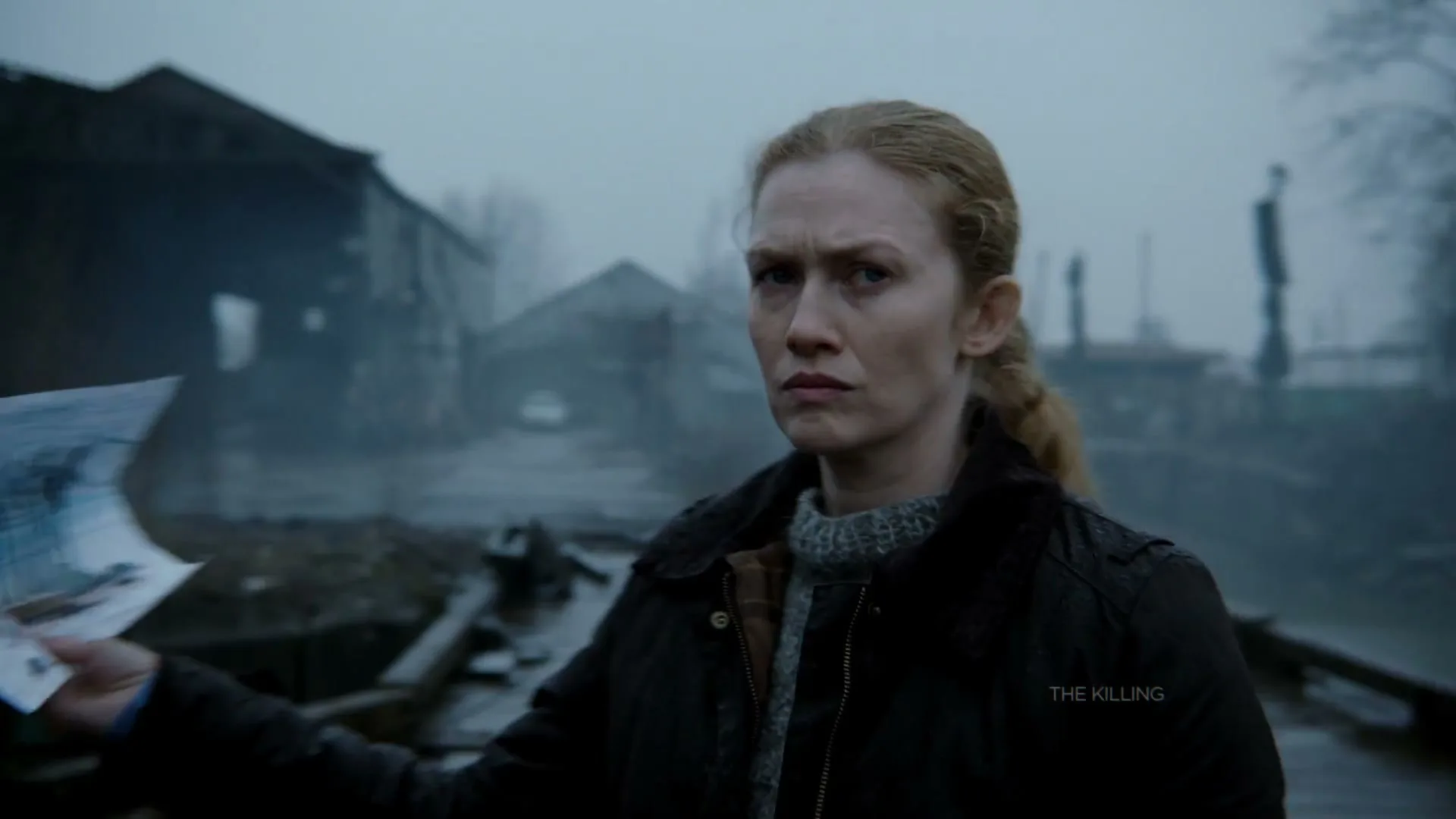 A woman with red hair in a dark jacket holds a drawing and looks intently at the camera in a foggy, industrial exterior setting.