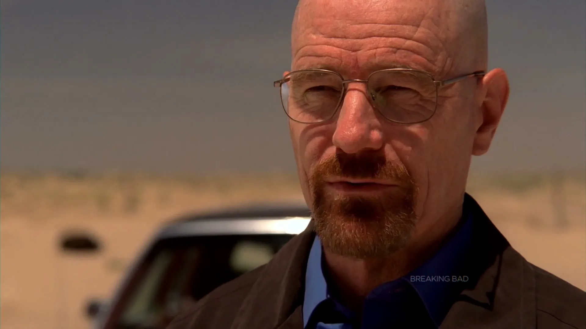 A bald man with glasses and a goatee looks directly at the camera in a desert landscape with a car in the background.