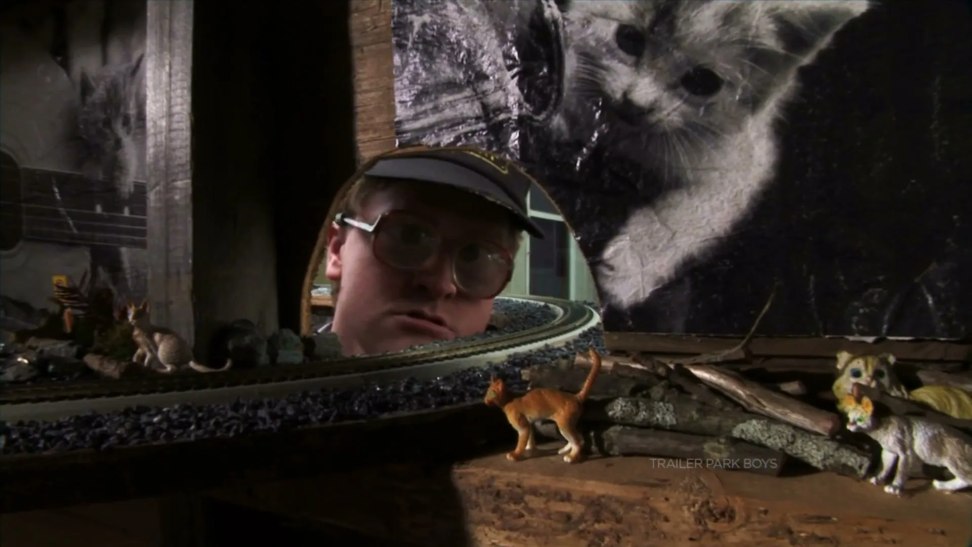 A man with glasses and a cap peeks through a hole in a wall, observing a miniature train set and cat figurines, with a large cat poster in the background.