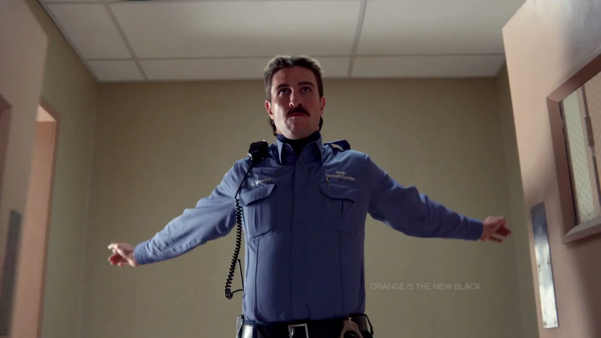 A male prison guard with a mustache stands in a hallway, holding two doors open with his arms outstretched, looking up.