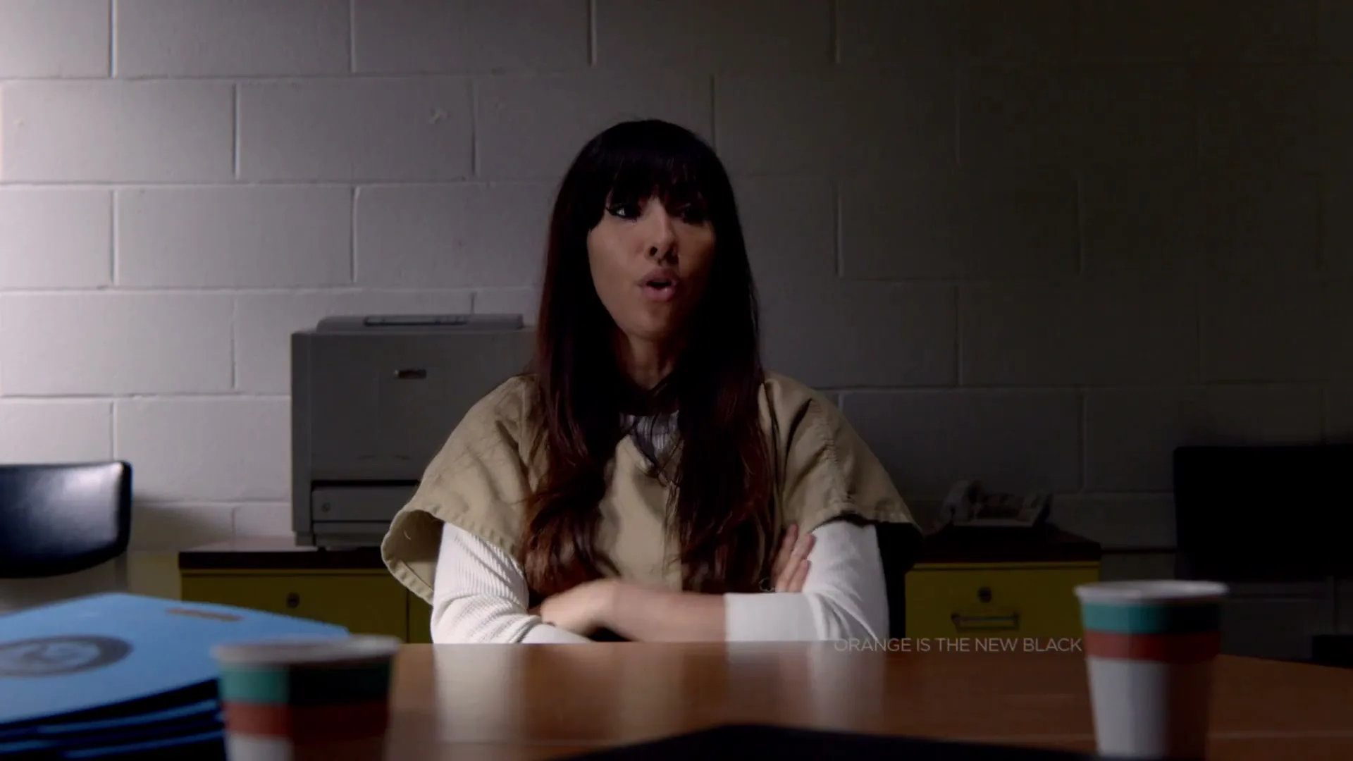 A woman with long dark hair, wearing a prison uniform, sits at a table in a dimly lit room, talking and gesturing.